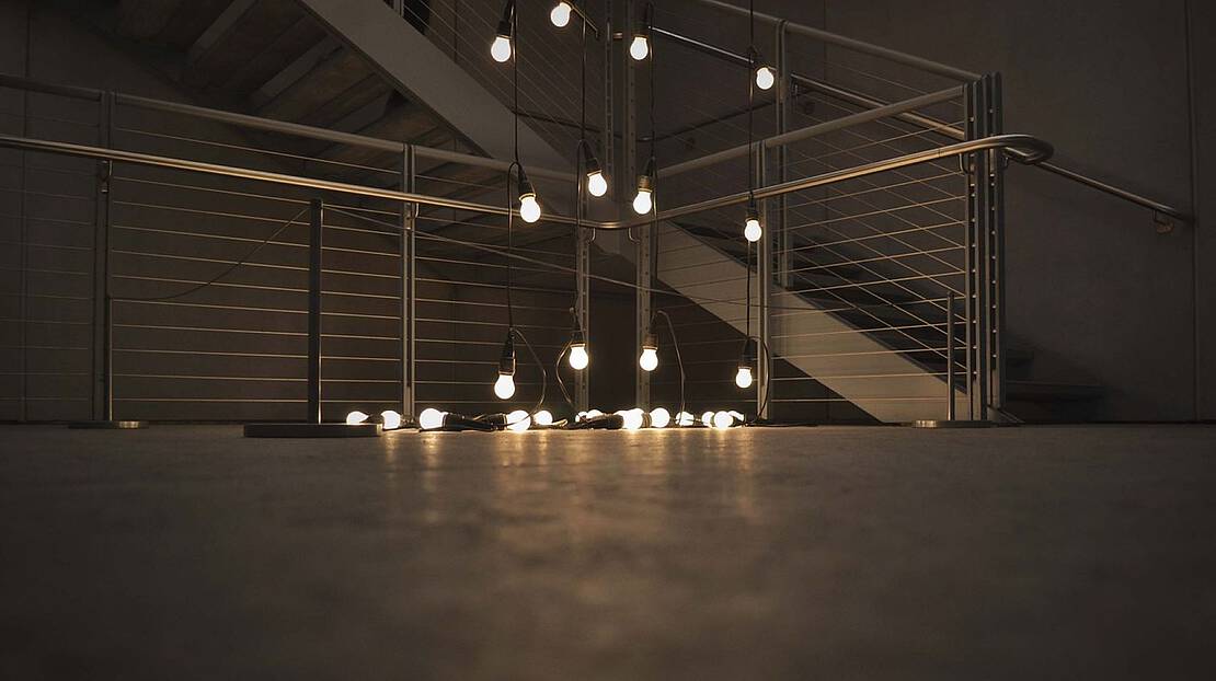 Eine angeschaltete Lichterkette mit Glühbirnen liegt in einem dunklen Treppenhaus.